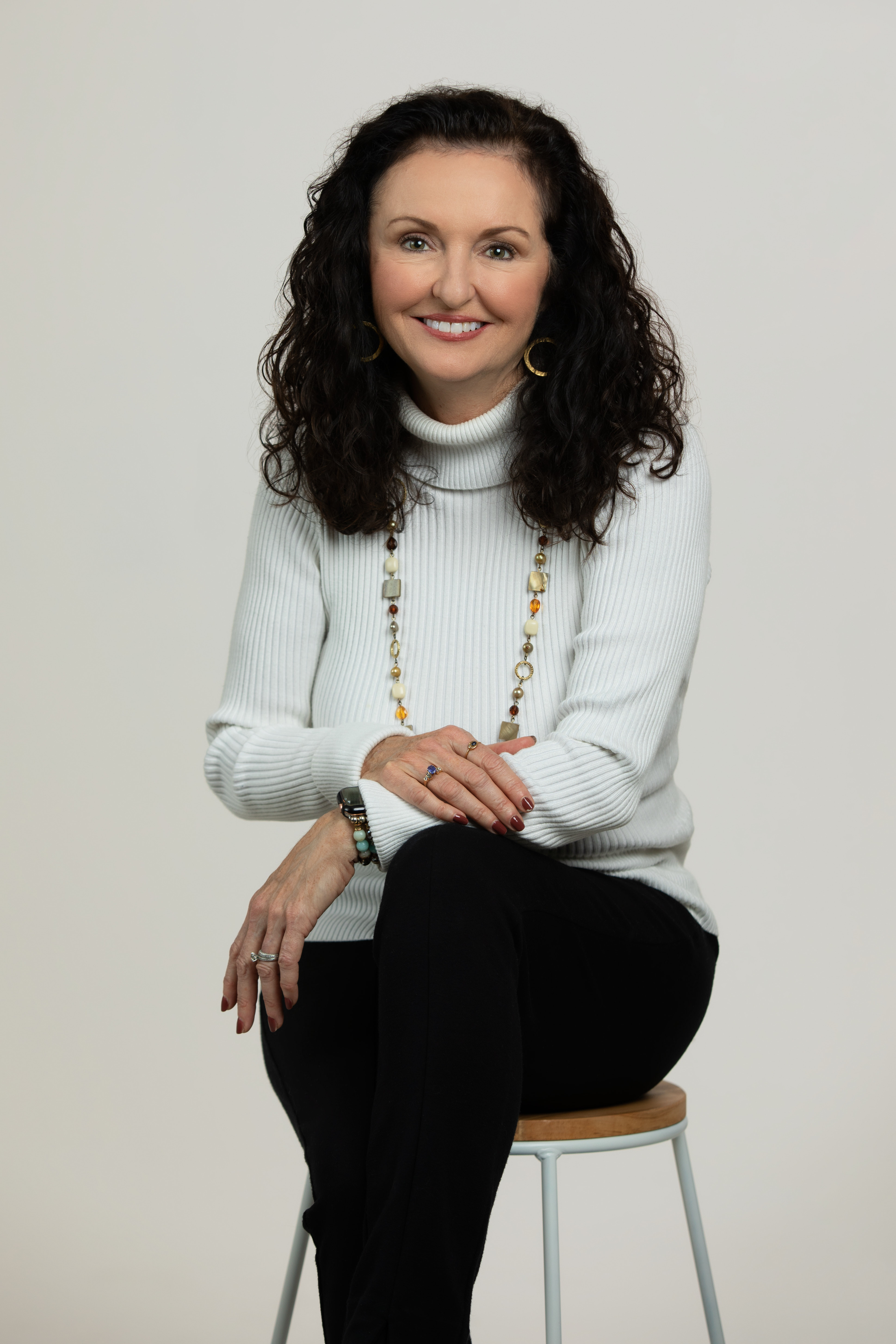 Dark haired woman sitting on a stool wearing white turtleneck.